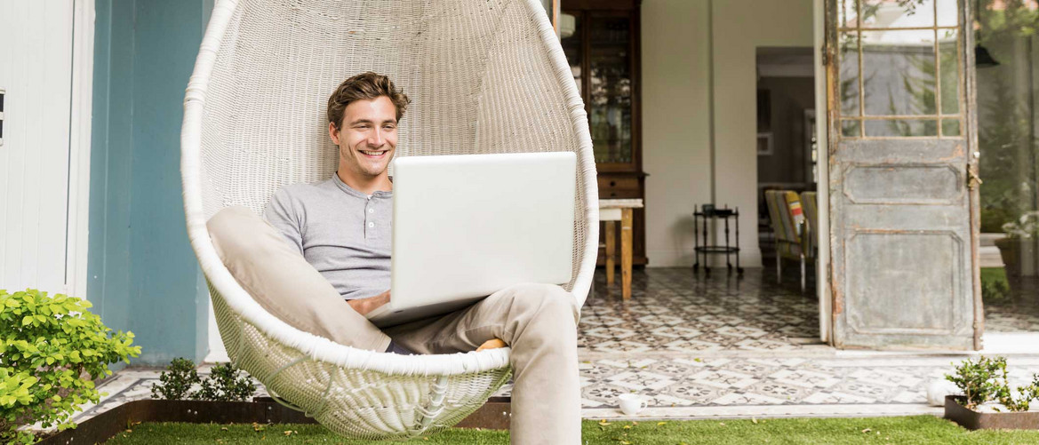 Glücklicher junger Mann mit Laptop in einem weißen Outdoor-Hängesessel.