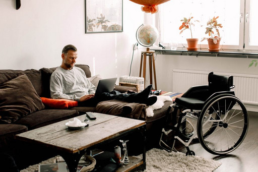 Mann sitzt im Wohnzimmer auf dem Sofa, vor dem ein Rollstuhl steht