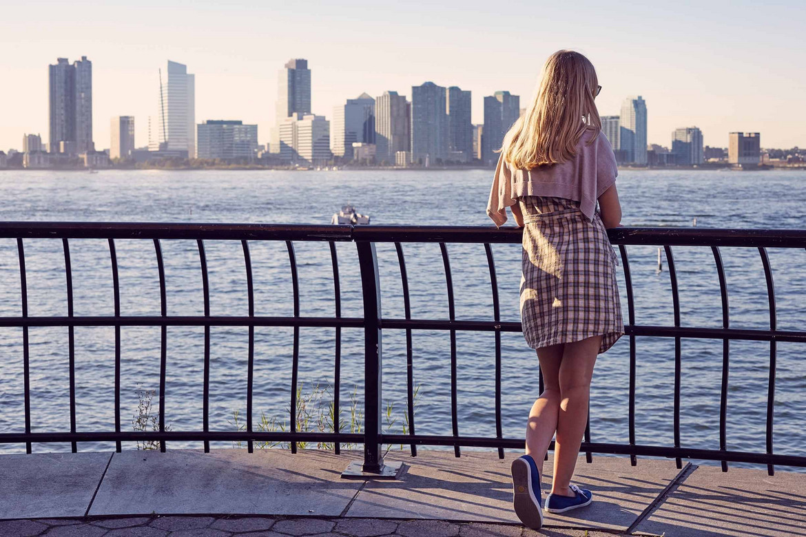 Frau mit Loafern steht am Ufer und schaut auf die gegenüberliegende Skyline