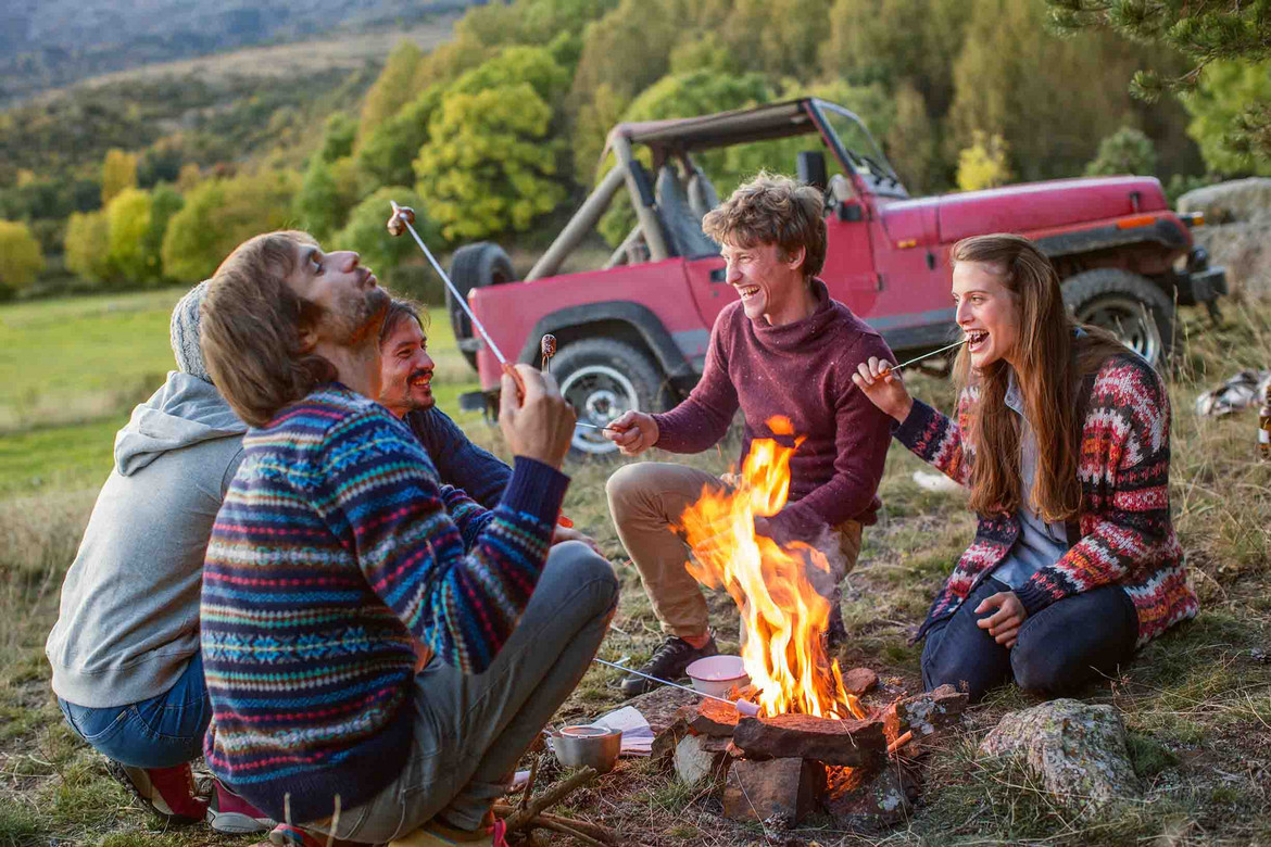 Vier Jugendliche sitzen um ein Lagerfeuer herum, im Hintergrund ein Geländewagen