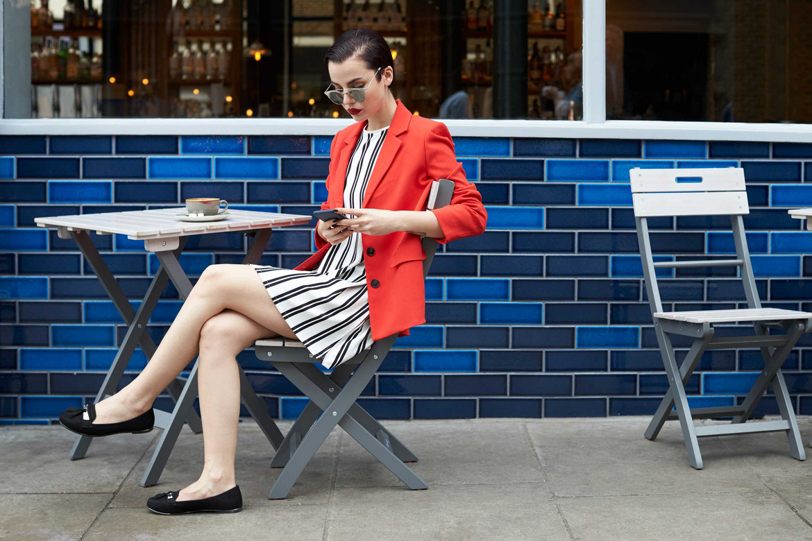 Frau mit rotem Blazer und Loafer sitzt in einem Straßencafé und schaut auf ihr Handy